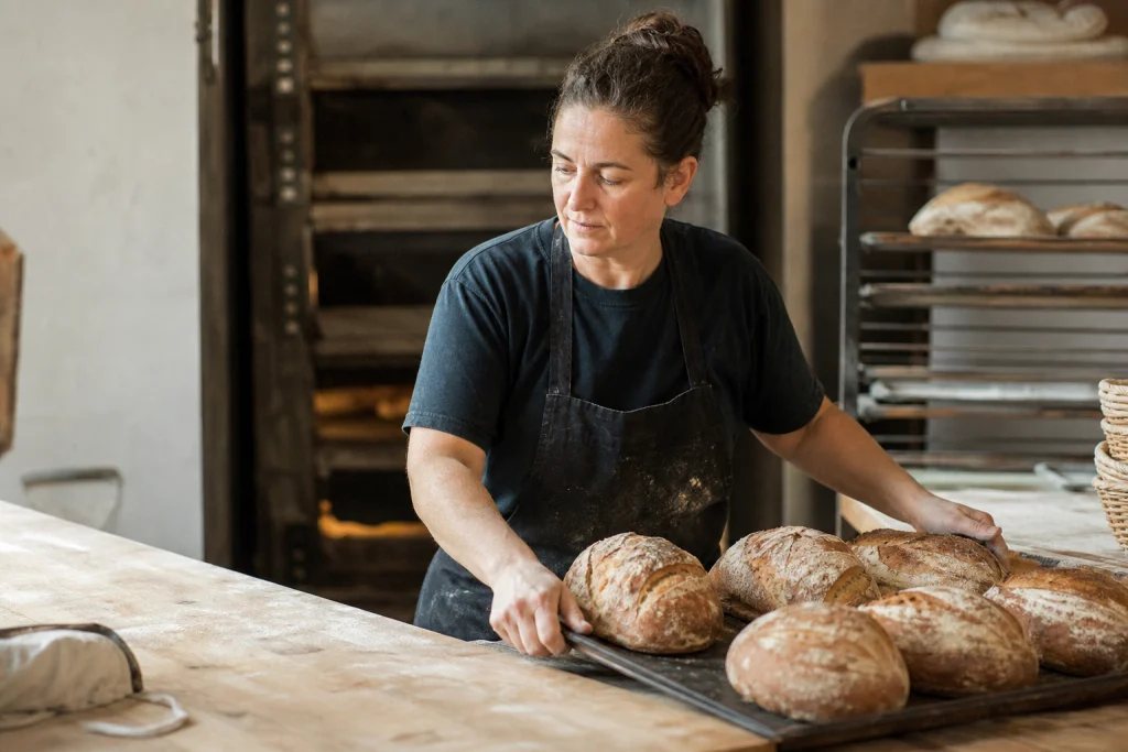 Woman baker arranging fresh bread in her small bakery, natural light, funding options for women-owned businesses